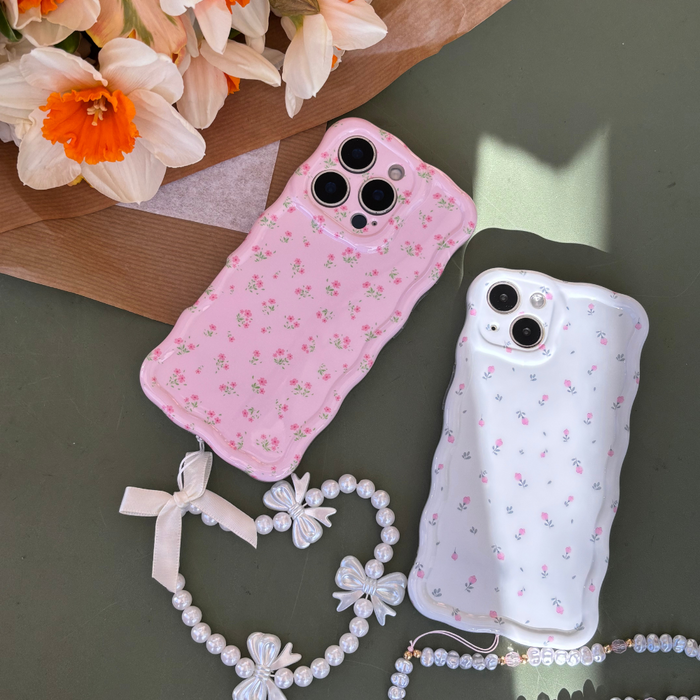 White Wavy Coconut Lane phone case with ditsy floral print, pearl and bow charms, placed by orange and white flowers.