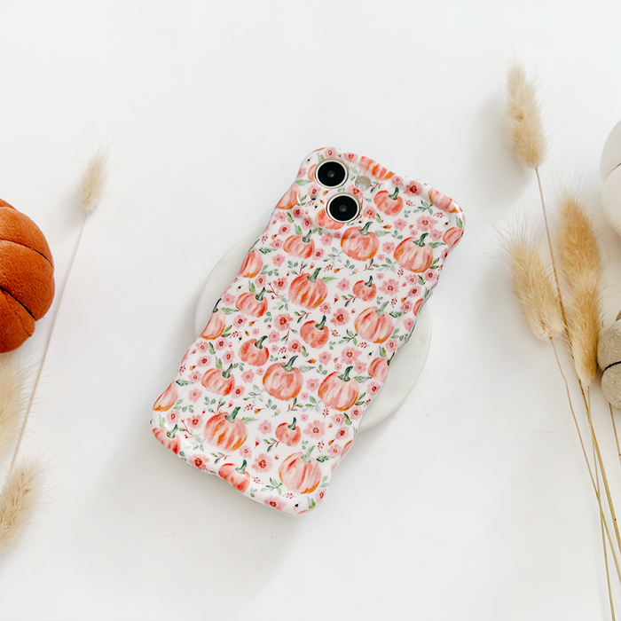 Wavy phone case with a ditsy pumpkin pattern on a white surface, next to dried grasses and plush pumpkins.