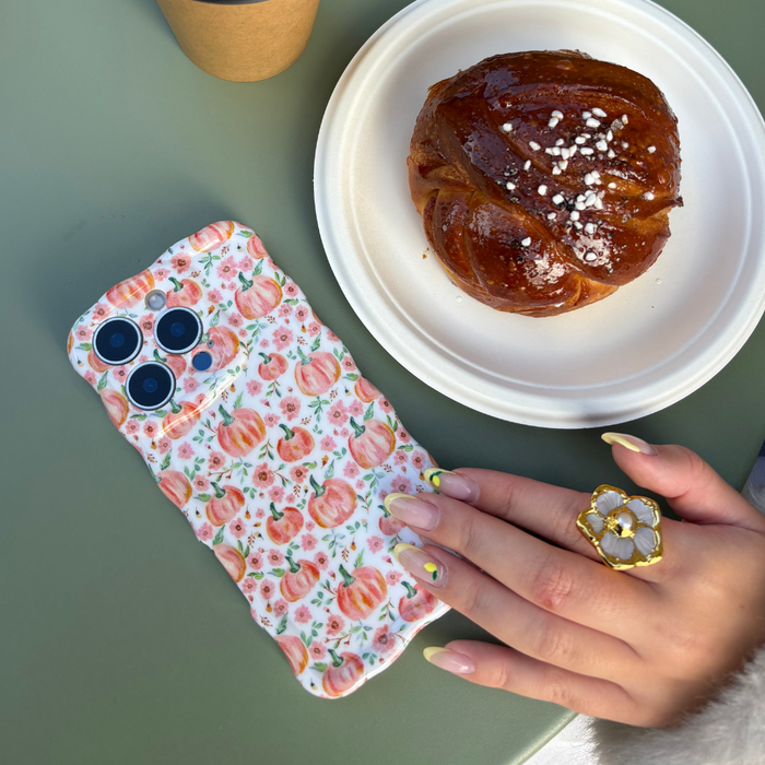 Hand with floral ring on Coconut Lane wavy phone case featuring a ditsy pumpkin pattern, near a glazed pastry on a plate.