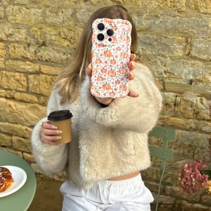 Woman in a beige fuzzy sweater holds coffee and a phone with a wavy case featuring an orange pumpkin pattern.