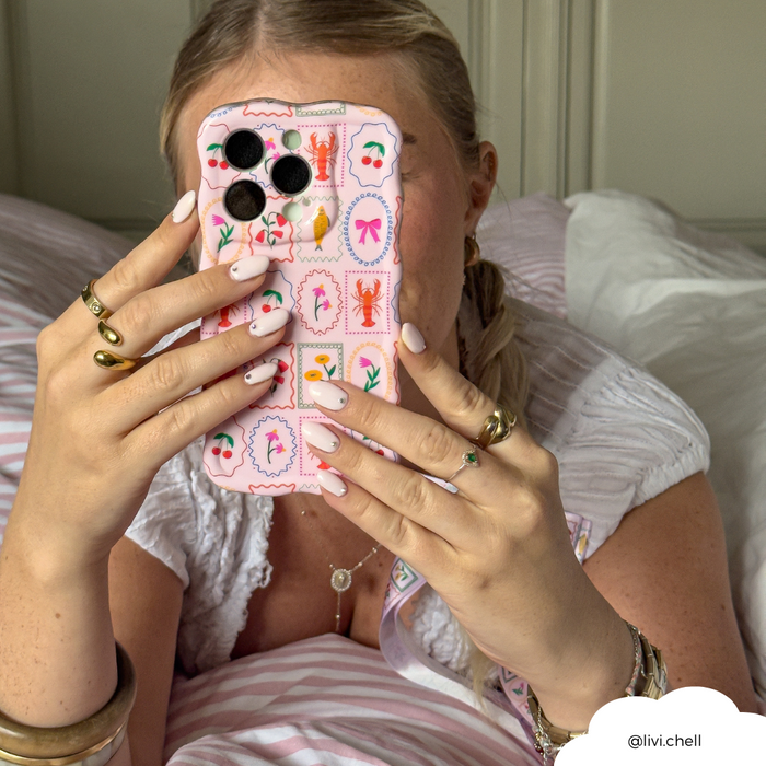 Woman with braided hair holds a wavy pastel phone case in front of her face while sitting on a bed.