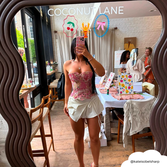 Woman in pink corset and white skirt holds a wavy Coconut Lane Dolce Vita phone case while taking a mirror selfie.