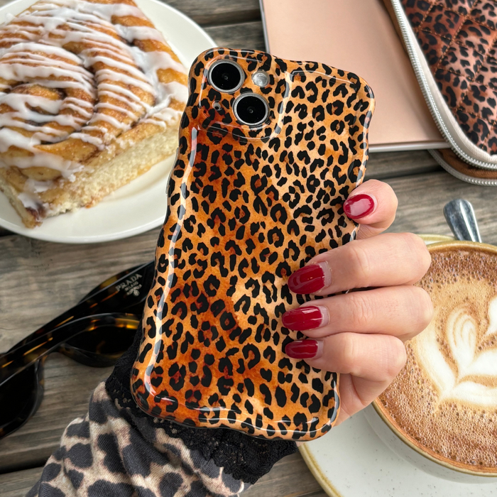 Red-nailed hand holds a wavy leopard-print phone case next to coffee, a pastry, sunglasses, and a pouch.