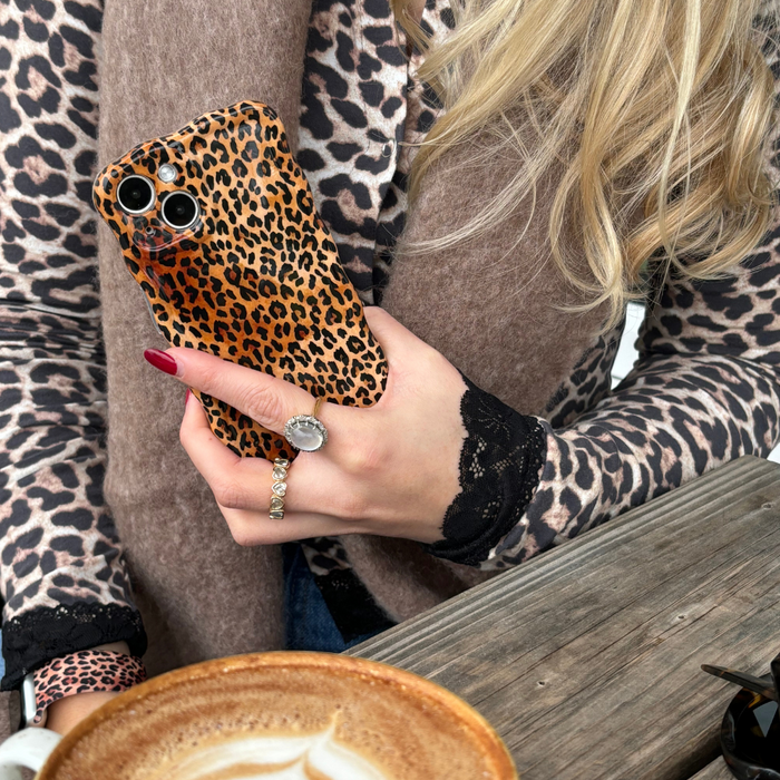 Woman holding a Coconut Lane leopard print wavy phone case, wearing a matching top and scarf at a wooden table.