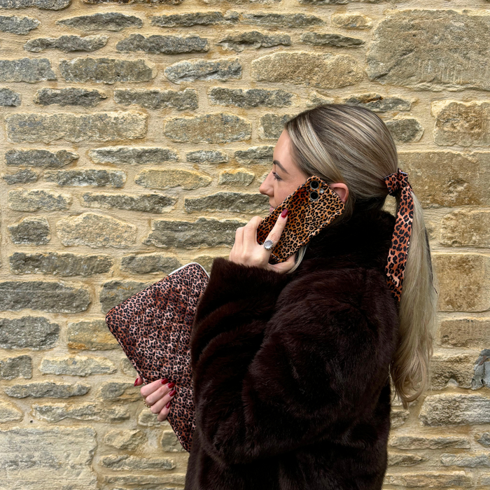 Woman in a dark fur coat holds a leopard print clutch, wearing a hair scarf and holding a wavy leopard print phone case.