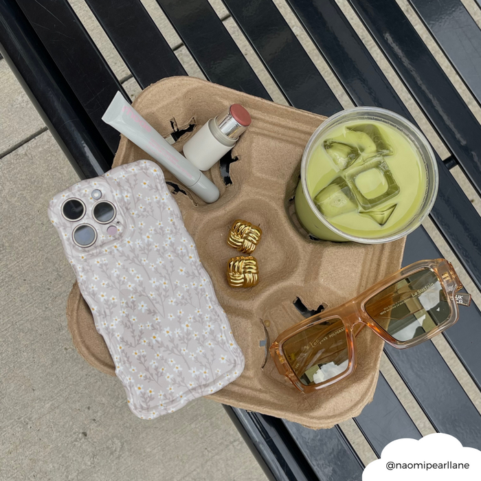 Tray with iced matcha, lipstick, gold rings, sunglasses, and a beige phone case with nude flower pattern.