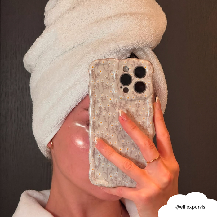 Person wearing a white towel turban and eye patches holds a phone with a nude floral wavy Coconut Lane case in a mirror selfie.