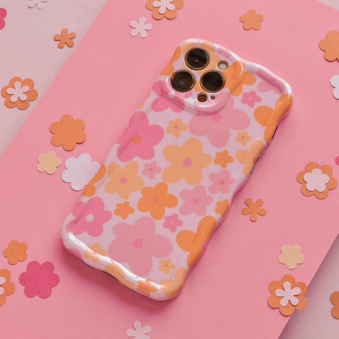 A Coconut Lane Wavy Phone Case - Pink & Orange Bloom lies on a pink surface scattered with small, flower-shaped cutouts in various shades of pink and orange. The case's extra protective design complements the cohesive, vibrant, and playful floral aesthetic.