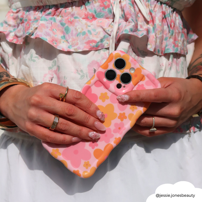 Woman holds Coconut Lane phone case in pink and orange wavy pattern, wearing a floral dress with manicured nails.