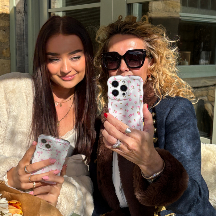 Two women smiling in sunlight, each holding a pink floral Coconut Lane wavy phone case.