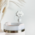 A close-up of a light pink tumbler with a silver accent. The tumbler features a white straw topped with Coconut Lane's adorable White Cloud Straw Cover accessory. In the background, there are blurred beige pampas grass against a light, neutral wall.